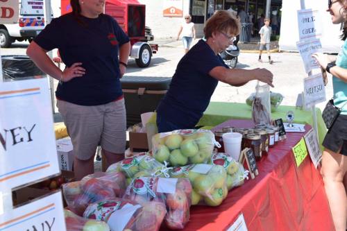 A vendor at a market displays fresh apples and jars of preserves while interacting with a customer.