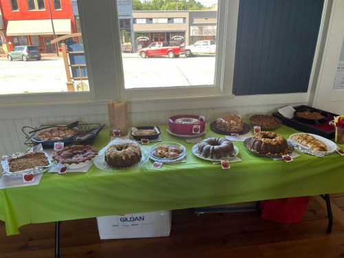 A table displays various baked goods, including cakes and cookies, with a green tablecloth and a street view in the background.