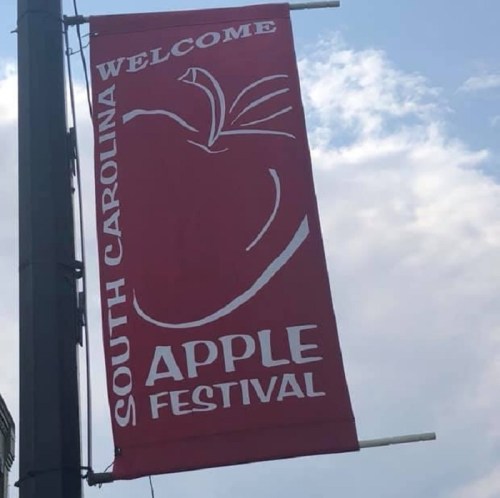 Red banner reading "South Carolina Welcome Apple Festival" with an apple graphic against a cloudy sky.