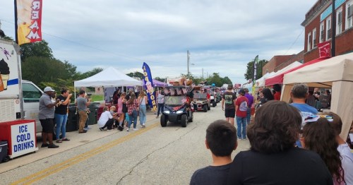 A parade with decorated golf carts moves down a street lined with vendors and spectators enjoying the event.