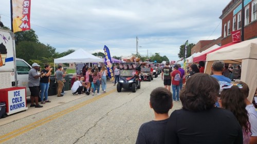 A parade with decorated golf carts moves down a street lined with food stalls and spectators.