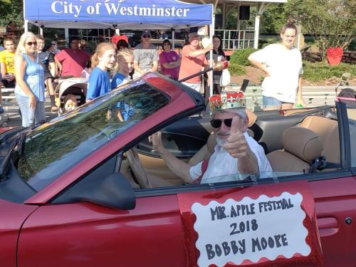 A man in a convertible car wearing a crown and pointing, with a sign reading "Mr. Apple Festival 2018 Bobby Moote."