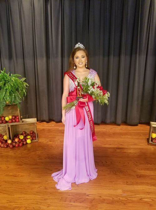 A young woman in a purple gown holds a trophy and flowers, wearing a tiara and sash, standing on a stage.