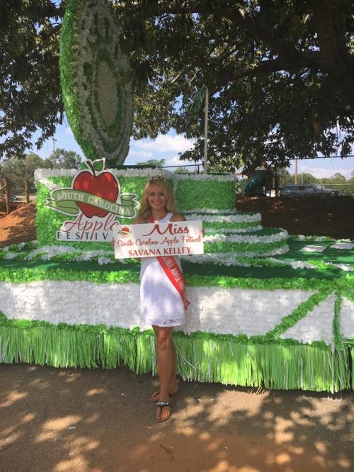 A young woman in a white dress holds a banner at the South Carolina Apple Festival, standing in front of a decorated float.