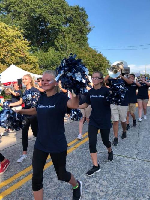 A marching band parades down a street, with members holding pom-poms and instruments, surrounded by trees and tents.