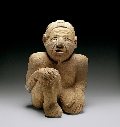 A seated, abstract sculpture of a human figure, carved from stone, with a contemplative expression and simplified features.