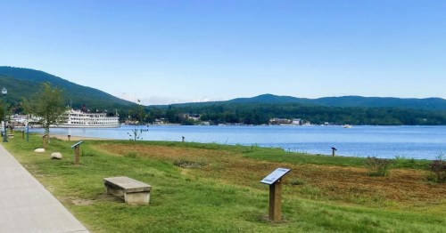 Views of Lake George framed by lush rolling hills in the distance, with a steamboat, and featuring clear blue waters.