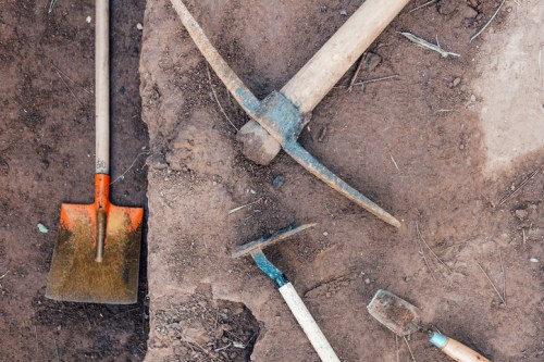 A flat lay of various gardening tools, including a shovel, pickaxe, and hoe, on brown soil.