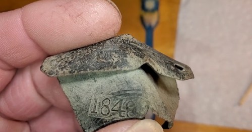 A close-up of a weathered metal object with the number "1849" engraved on it, held between fingers.