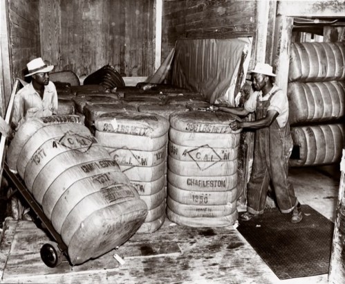 Two men in hats handle large barrels in a warehouse, surrounded by stacked bales and wooden walls.
