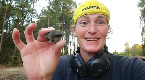 A smiling person wearing a yellow cap and headphones holds up a small rock in a forested area.