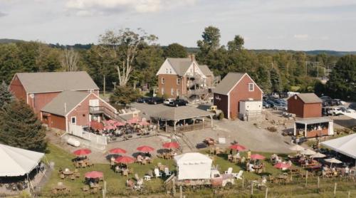 Aerial view of a rustic farm with red buildings, outdoor seating, and umbrellas, surrounded by greenery and parked cars.