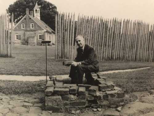 A man kneels by a stone foundation with a historic building and wooden fence in the background.