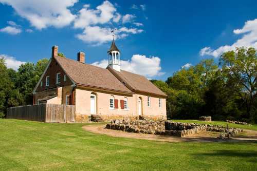 Historic building with a bell tower, surrounded by green grass and trees under a blue sky with fluffy clouds.