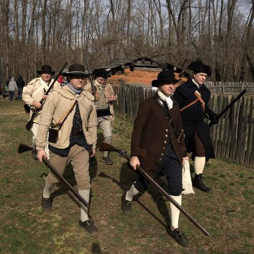 A group of men in historical military attire march with rifles, surrounded by trees and a rustic building.