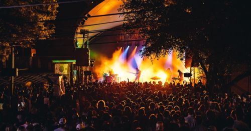 A large crowd enjoys a live music performance under colorful stage lights at an outdoor venue.