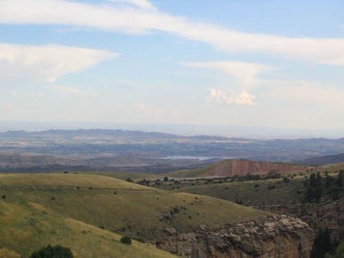 A panoramic view of rolling hills and distant mountains under a blue sky with scattered clouds.