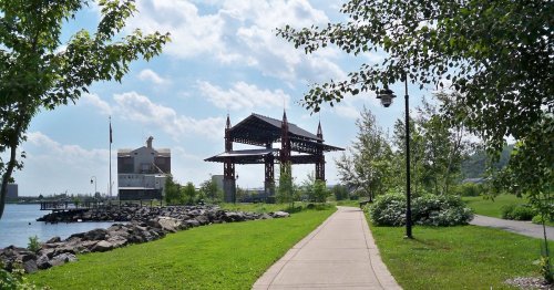 A scenic park path alongside a lake, featuring greenery, a pavilion, and a clear blue sky with scattered clouds.