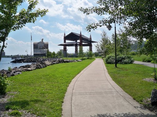 A paved path leads along a grassy area by the water, with trees and a large structure in the background under a blue sky.
