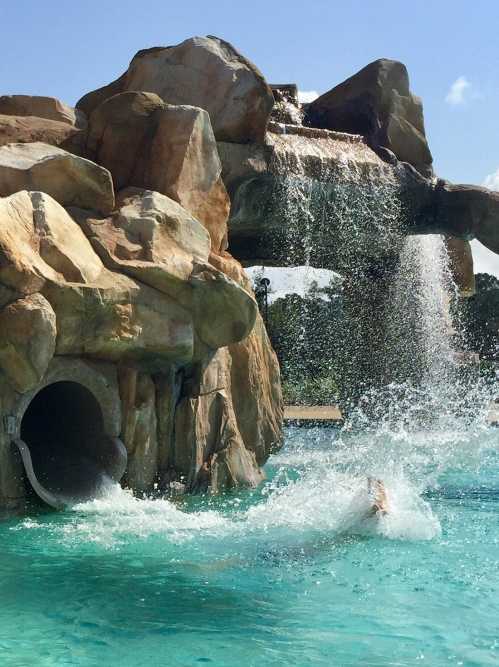 A water slide with a rocky structure, cascading water, and a splash in a bright blue pool under a clear sky.