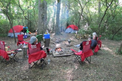 A campsite with two tents, a campfire, and people sitting in chairs among trees in a wooded area.