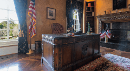 A classic office with a wooden desk, American flags, and elegant decor, featuring a large window and a fireplace.