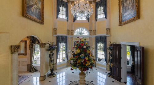 Luxurious foyer with a grand chandelier, floral arrangement, and elegant decor, featuring large windows and ornate details.