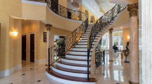 Elegant spiral staircase with ornate iron railing in a bright, spacious foyer featuring marble floors and warm yellow walls.