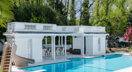 A white poolside structure with columns, a slide, and seating area beside a clear blue pool surrounded by greenery.