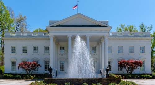 A grand white mansion with columns, a fountain in front, and an American flag flying on a clear blue day.