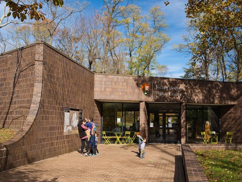 A modern visitor center surrounded by trees, with people outside and bright green chairs on the patio.
