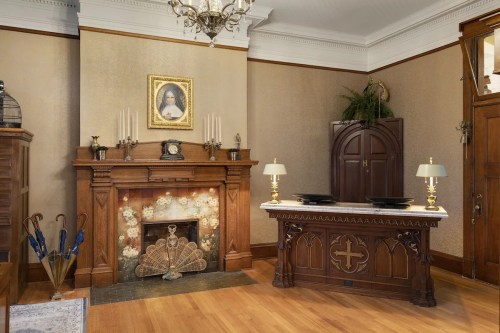 A vintage room featuring a wooden fireplace, ornate decor, a portrait, and a table with lamps.