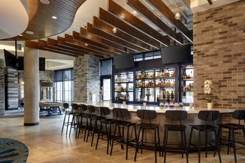 Modern bar interior with a sleek counter, high stools, and a well-stocked shelf of liquor behind it.