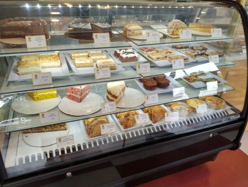 A display case filled with various desserts, including cakes, pastries, and slices, each labeled with names.