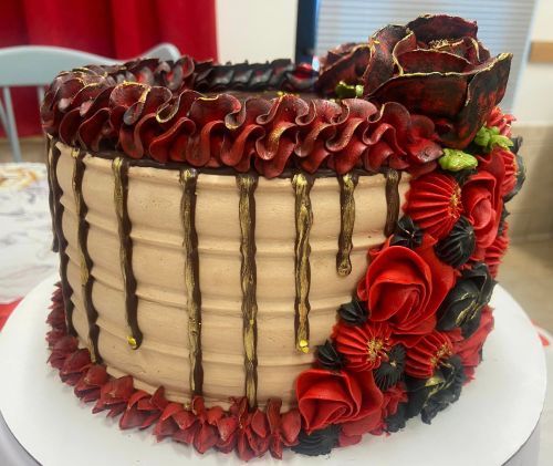 A beautifully decorated cake with red and black frosting, featuring roses and ruffled edges, topped with gold accents.