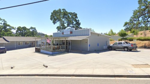 A small building with a porch, surrounded by trees and a parking area, under a clear blue sky.