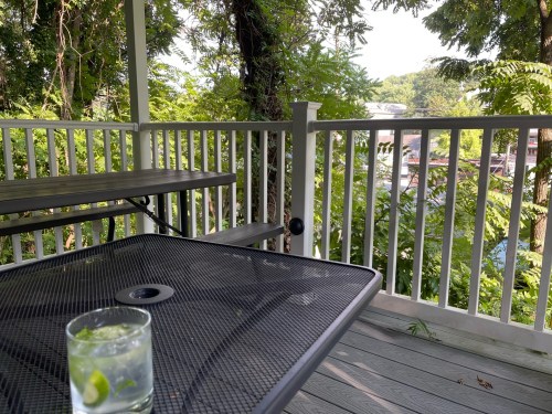 A table on a porch with a drink, surrounded by greenery and a view of distant trees and buildings.
