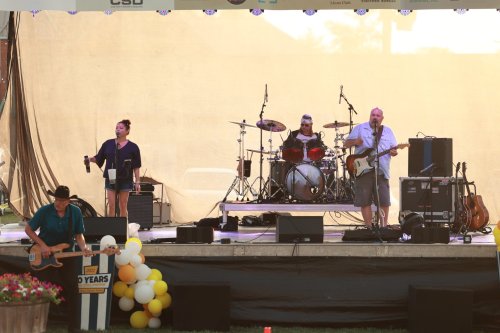 A band performs on stage with a singer, drummer, and guitarist, set against a backdrop of balloons and equipment.