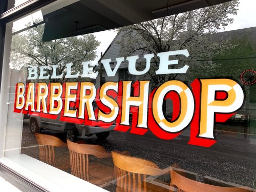 Colorful sign reading "Bellevue Barbershop" on a shop window, reflecting trees outside.
