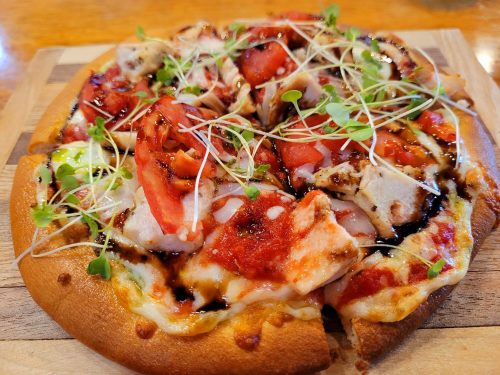 A close-up of a pizza topped with chicken, tomatoes, sprouts, and drizzled with sauce on a wooden board.
