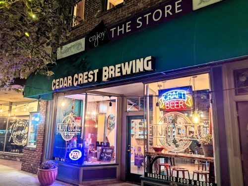 Exterior of CedarCrest Brewing with neon signs, showcasing "Craft Beer" and "Enjoy Local" under a vibrant storefront.