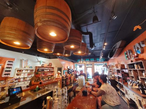 A cozy shop interior with wooden barrels hanging from the ceiling, a bar, and customers enjoying drinks at the counter.