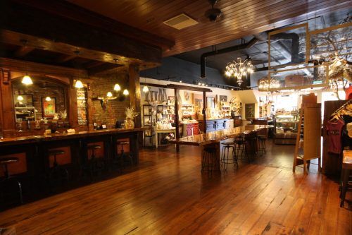 A cozy interior of a shop with wooden floors, a bar area, and shelves filled with various products. Warm lighting enhances the ambiance.