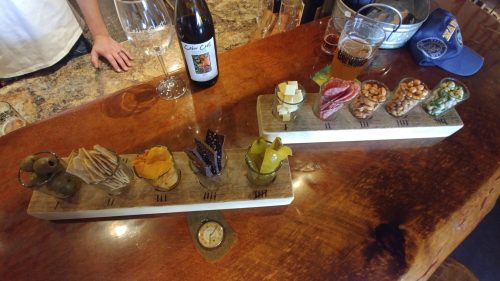 A wooden platter with various snacks: olives, cheese, meats, nuts, and chocolates, alongside wine and beer glasses.