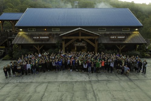 A large group of people gathered in front of a rustic building with a blue roof, surrounded by trees and mountains.