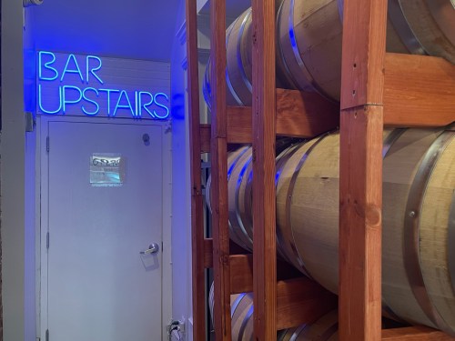 A wooden barrel rack beside a door with a glowing "BAR UPSTAIRS" sign in blue neon light.