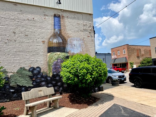 A mural of a wine bottle and grapes on a brick wall, with a bench and cars nearby under a partly cloudy sky.