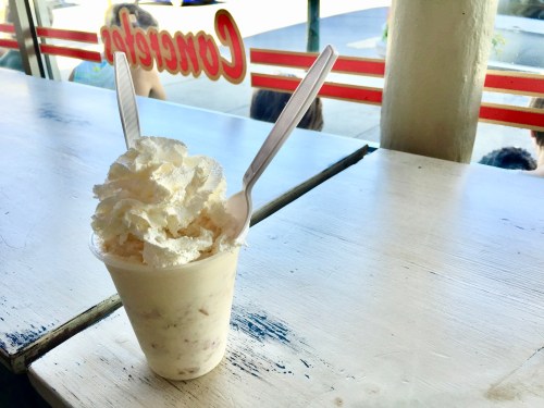 A cup of ice cream topped with whipped cream, placed on a table with a window view in the background.