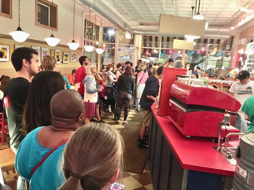 A crowded restaurant interior with people waiting in line at the counter, colorful decor, and warm lighting.