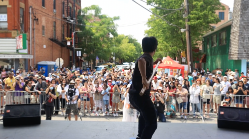 A performer dances on stage in front of a large crowd at an outdoor event, with trees and buildings in the background.